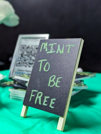 A small chalkboard sign with the words 'MINT TO BE FREE' written in green chalk. The sign is placed on a green tablecloth with some blurred items, including a QR code, in the background.