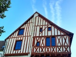 A traditional half-timbered house with a steeply pitched roof. The structure features exposed wooden beams creating geometric patterns, filled with white stucco. There are several dark-framed windows, and part of a tree with green leaves is visible on the left side against a clear blue sky with a few wisps of cloud.
