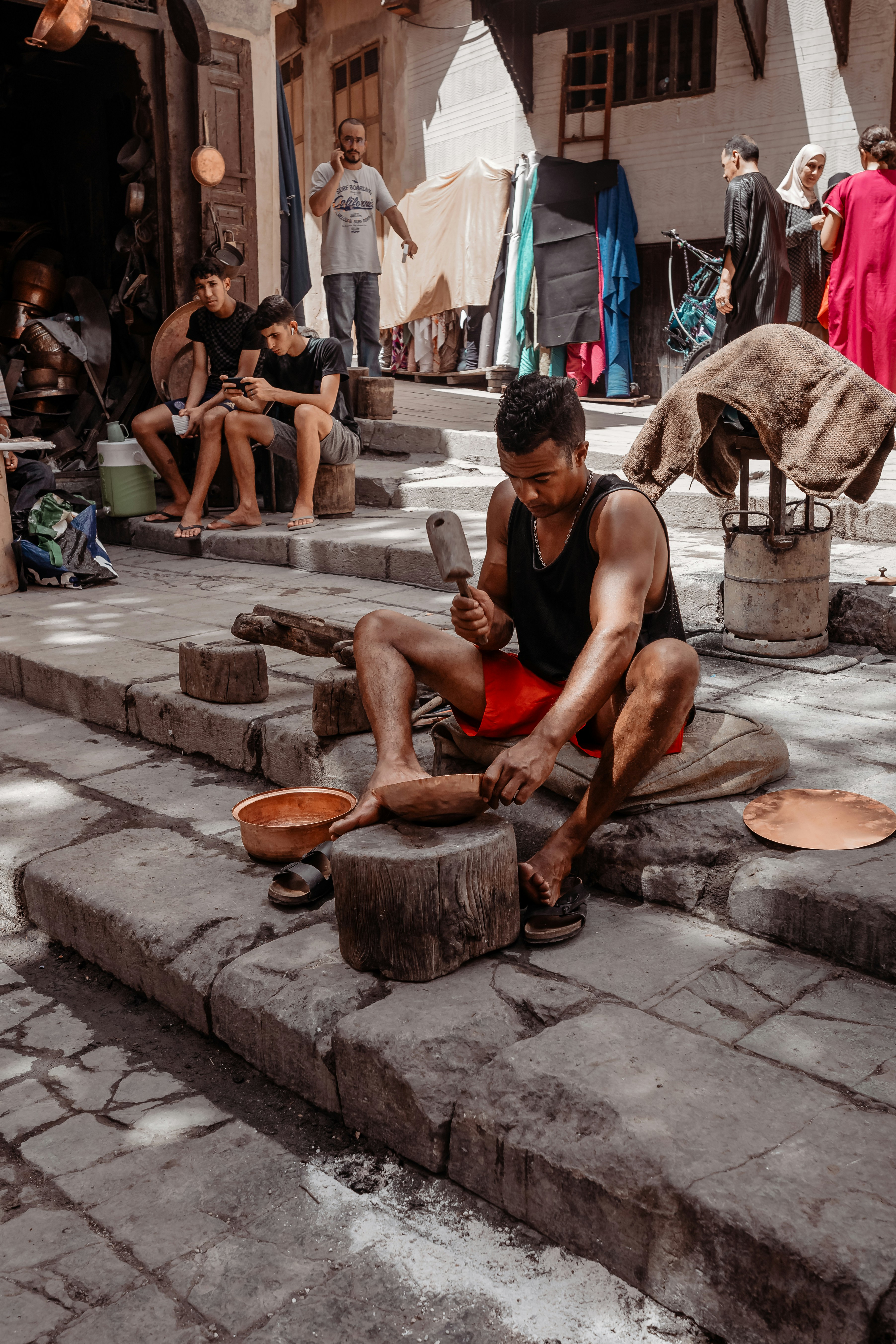 A person is working with metal tools on stone steps, likely crafting or shaping a metal object. Several other individuals are present, some seated and possibly using mobile devices. The background shows a street or market setting with a variety of fabrics and goods hanging and displayed, and people walking by.