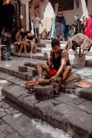 A person is working with metal tools on stone steps, likely crafting or shaping a metal object. Several other individuals are present, some seated and possibly using mobile devices. The background shows a street or market setting with a variety of fabrics and goods hanging and displayed, and people walking by.