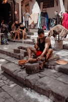 A person is working with metal tools on stone steps, likely crafting or shaping a metal object. Several other individuals are present, some seated and possibly using mobile devices. The background shows a street or market setting with a variety of fabrics and goods hanging and displayed, and people walking by.