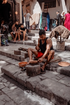 A person is working with metal tools on stone steps, likely crafting or shaping a metal object. Several other individuals are present, some seated and possibly using mobile devices. The background shows a street or market setting with a variety of fabrics and goods hanging and displayed, and people walking by.