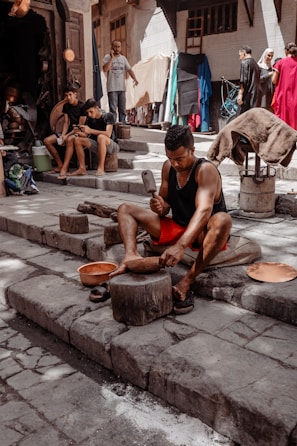 A person is working with metal tools on stone steps, likely crafting or shaping a metal object. Several other individuals are present, some seated and possibly using mobile devices. The background shows a street or market setting with a variety of fabrics and goods hanging and displayed, and people walking by.