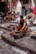 A person is working with metal tools on stone steps, likely crafting or shaping a metal object. Several other individuals are present, some seated and possibly using mobile devices. The background shows a street or market setting with a variety of fabrics and goods hanging and displayed, and people walking by.