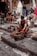 A person is working with metal tools on stone steps, likely crafting or shaping a metal object. Several other individuals are present, some seated and possibly using mobile devices. The background shows a street or market setting with a variety of fabrics and goods hanging and displayed, and people walking by.