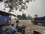 A rural roadside scene with a makeshift food stall on the left, featuring a table with pots and condiments under a blue tarpaulin. A motorcycle rests nearby, while a yellow truck is parked on the right side. The background shows trees, including a few palm trees, and a large building, suggesting a rural transport hub or market area. The ground is dusty and littered with debris, and the sky appears overcast.
