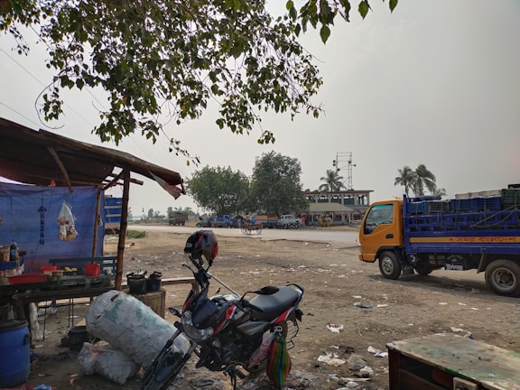 A rural roadside scene with a makeshift food stall on the left, featuring a table with pots and condiments under a blue tarpaulin. A motorcycle rests nearby, while a yellow truck is parked on the right side. The background shows trees, including a few palm trees, and a large building, suggesting a rural transport hub or market area. The ground is dusty and littered with debris, and the sky appears overcast.