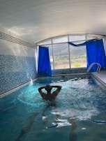 An indoor swimming pool area with blue mosaic-tiled walls and floor. A person is standing in the pool, enjoying the water, with hands clasped behind the head. Large windows with deep blue curtains are open, revealing a view of trees and mountains outside. The ceiling is arched and white, contributing to a bright atmosphere.