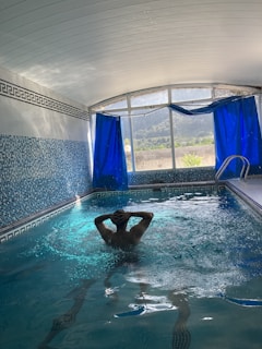 An indoor swimming pool area with blue mosaic-tiled walls and floor. A person is standing in the pool, enjoying the water, with hands clasped behind the head. Large windows with deep blue curtains are open, revealing a view of trees and mountains outside. The ceiling is arched and white, contributing to a bright atmosphere.