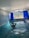 An indoor swimming pool area with blue mosaic-tiled walls and floor. A person is standing in the pool, enjoying the water, with hands clasped behind the head. Large windows with deep blue curtains are open, revealing a view of trees and mountains outside. The ceiling is arched and white, contributing to a bright atmosphere.