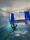 An indoor swimming pool area with blue mosaic-tiled walls and floor. A person is standing in the pool, enjoying the water, with hands clasped behind the head. Large windows with deep blue curtains are open, revealing a view of trees and mountains outside. The ceiling is arched and white, contributing to a bright atmosphere.