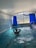 An indoor swimming pool area with blue mosaic-tiled walls and floor. A person is standing in the pool, enjoying the water, with hands clasped behind the head. Large windows with deep blue curtains are open, revealing a view of trees and mountains outside. The ceiling is arched and white, contributing to a bright atmosphere.