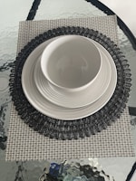 A dining setup featuring a white bowl placed on a stack of white plates. The stack is on a dark intricately patterned round placemat, which is on top of a square, textured beige mat. The entire arrangement rests on a glass table with a rippled surface.