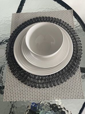 A dining setup featuring a white bowl placed on a stack of white plates. The stack is on a dark intricately patterned round placemat, which is on top of a square, textured beige mat. The entire arrangement rests on a glass table with a rippled surface.
