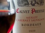 Close-up of a wine bottle label showing the name Calvet Prestige, Merlot, Cabernet Sauvignon, Bordeaux, and the year 2020. A glass of red wine is partially visible to the left.