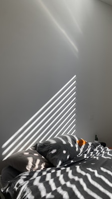 Sunlight filters through blinds, casting striped shadows across a neatly made bed with pillows. The room appears minimalist with a focus on natural light and simplicity.
