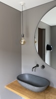 A modern bathroom interior with a minimalist design, featuring a round mirror mounted on a gray wall. Below the mirror, a sleek, black sink is set on a wooden countertop. A wall-mounted faucet is positioned above the sink. To the left, a hanging light bulb with a visible filament dangles from the ceiling.