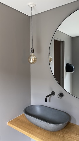 A modern bathroom interior with a minimalist design, featuring a round mirror mounted on a gray wall. Below the mirror, a sleek, black sink is set on a wooden countertop. A wall-mounted faucet is positioned above the sink. To the left, a hanging light bulb with a visible filament dangles from the ceiling.