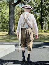 A person dressed in historical clothing, including a tricorn hat, loose white shirt, and brown knee-length trousers, walks on a paved path in a wooded area. The individual carries a folding chair in one hand, with suspenders visible across their back.