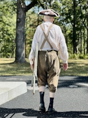 A person dressed in historical clothing, including a tricorn hat, loose white shirt, and brown knee-length trousers, walks on a paved path in a wooded area. The individual carries a folding chair in one hand, with suspenders visible across their back.