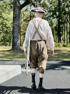 A person dressed in historical clothing, including a tricorn hat, loose white shirt, and brown knee-length trousers, walks on a paved path in a wooded area. The individual carries a folding chair in one hand, with suspenders visible across their back.