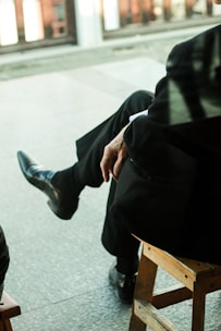 A person in formal attire sits on a wooden chair with their legs crossed, wearing black leather shoes and a dark suit. The background is blurred, indicating an indoor or outdoor setting with soft natural light.
