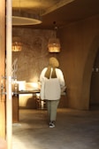 A person wearing a hijab is walking away in an interior space with earthy tones. The wall features a large mural with ancient Egyptian motifs under warm, ambient lighting. There are rustic lamp shades and a wooden table with some objects on it.