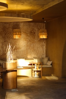 A cozy, warmly lit interior featuring a rustic wooden table and chair set against a wall with intricate Egyptian-style carvings. The lighting creates a serene atmosphere, with lampshades made of natural fibers adding an organic touch. Cushions adorn a built-in seating area, and decorative branches are arranged in a vase.