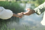 Two hands reaching out towards each other against a serene natural background with water and greenery. One person is wearing a beige long-sleeved shirt, and the other appears to have a bracelet on their wrist.