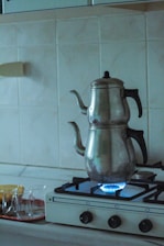 A metal teapot sits on a gas stove with a visible blue flame. The stovetop is white with black knobs, set against a tiled kitchen backsplash. Nearby, there is a tray with several small tea glasses.