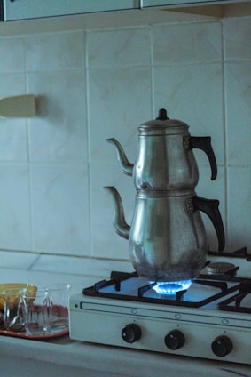 A metal teapot sits on a gas stove with a visible blue flame. The stovetop is white with black knobs, set against a tiled kitchen backsplash. Nearby, there is a tray with several small tea glasses.