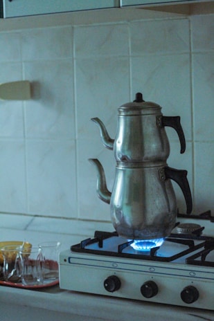 A metal teapot sits on a gas stove with a visible blue flame. The stovetop is white with black knobs, set against a tiled kitchen backsplash. Nearby, there is a tray with several small tea glasses.