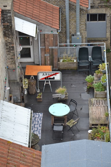 An urban rooftop patio contains a table with two chairs, a sign with an overlaid silhouette of a city skyline, various potted plants, wooden pallets, a patterned rug, and two stacked chairs. The area is surrounded by brick walls and grey pipes, with string lights adding a decorative touch.