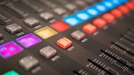 Close-up view of an audio mixing console with various colorful illuminated buttons and sliders. The console features sections labeled with terms like 'mute' and 'solo', displaying vibrant lights in shades of blue, red, and purple.