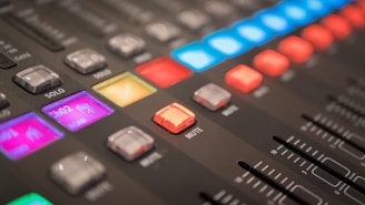 Close-up view of an audio mixing console with various colorful illuminated buttons and sliders. The console features sections labeled with terms like 'mute' and 'solo', displaying vibrant lights in shades of blue, red, and purple.