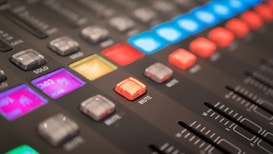 Close-up view of an audio mixing console with various colorful illuminated buttons and sliders. The console features sections labeled with terms like 'mute' and 'solo', displaying vibrant lights in shades of blue, red, and purple.