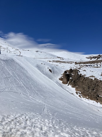 A snow-covered mountainous landscape with a clear blue sky. Ski tracks are visible on the slope, and there are rocky outcrops on the right side. A few structures for ski lifts can be seen in the distance.