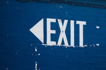 A painted exit sign on a blue brick wall, featuring a white arrow pointing left and the word 'EXIT' in bold white letters.