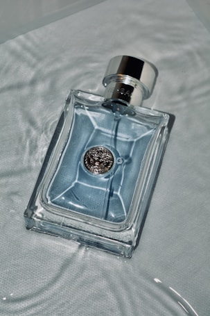 A clear glass bottle lies on a textured surface. The bottle contains a light blue liquid and has a metallic emblem on its front, with a shiny silver cap. Soft lighting creates subtle reflections on the liquid and glass.