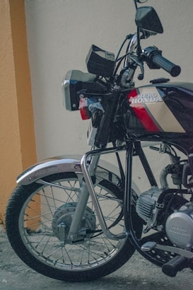 A close-up view of a motorcycle parked next to a wall. The motorcycle has shiny metal parts, a visible front wheel with spokes, and handlebars with a rearview mirror. The brand name 'Hero Honda' is prominently displayed on the fuel tank.