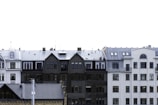 A row of European-style residential buildings with varied architectural designs. The buildings feature multiple stories with a mix of gabled and flat roofs. Windows vary in size and shape, with some rounded at the tops. The color palette includes shades of white, gray, and black. The sky is bright and clear, suggesting it could be a cold season due to the frosty appearance on some roofs.