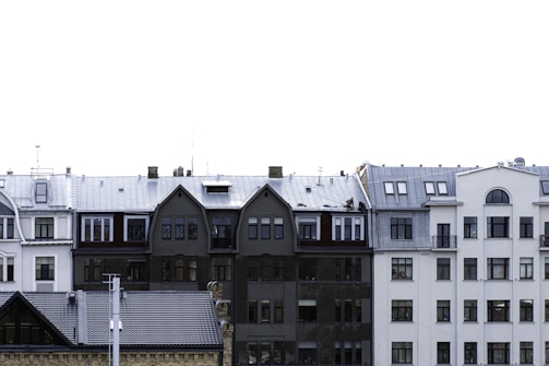 A row of European-style residential buildings with varied architectural designs. The buildings feature multiple stories with a mix of gabled and flat roofs. Windows vary in size and shape, with some rounded at the tops. The color palette includes shades of white, gray, and black. The sky is bright and clear, suggesting it could be a cold season due to the frosty appearance on some roofs.