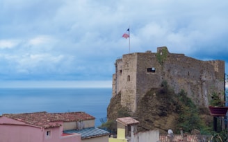 A weathered stone fortress with a flag on top overlooks a vast body of water under a cloudy sky. There are terracotta-roofed buildings in the foreground, nestled on a hillside adjacent to the fortress.