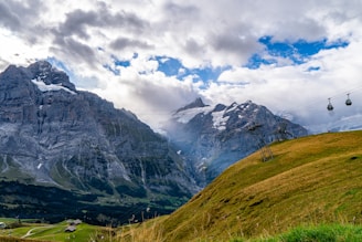 A scenic mountain landscape with rugged peaks partly covered in snow under a sky filled with dramatic clouds. In the foreground, grassy hills are traversed by a cable car system with gondolas suspended on wires, offering a breathtaking view.