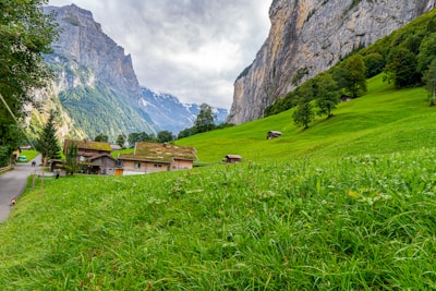 A serene alpine landscape features lush green meadows with scattered houses and a towering rocky mountain in the background. The sky is partly cloudy, creating a tranquil yet dramatic atmosphere.