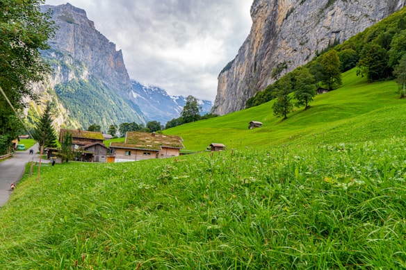 A serene alpine landscape features lush green meadows with scattered houses and a towering rocky mountain in the background. The sky is partly cloudy, creating a tranquil yet dramatic atmosphere.