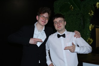 Two young men are posing together, dressed formally in black and white attire. They are smiling and appear to be having a good time, with one man wearing glasses. Both are pointing their fingers in a playful manner, standing against a dark backdrop with some greenery and decorative elements.