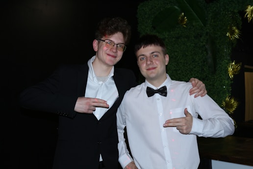 Two young men are posing together, dressed formally in black and white attire. They are smiling and appear to be having a good time, with one man wearing glasses. Both are pointing their fingers in a playful manner, standing against a dark backdrop with some greenery and decorative elements.