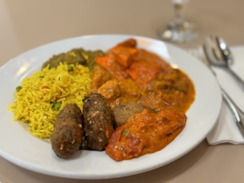 A plate of Indian cuisine featuring vibrant yellow rice with peas, assorted curries with rich red and brown gravies, and two brown kebabs. The dish is served on a white plate alongside cutlery on a neutral tablecloth background.