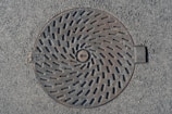 A round metal manhole cover is set in asphalt. The cover features a spiral pattern of slots and has text engraved on it. The surface shows signs of wear with a slightly weathered appearance.